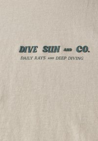 Světlý béžový textil s texturovaným, žebrovaným designem. Tmavě tyrkysový text zní: "DIVE SUN and CO. DAILY RAYS and DEEP DIVING."