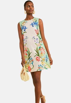 Moteris dėvi be rankovių gėlėtą suknelę pastelinių atspalvių, laiko pintinę rankinę, stovi susikryžiavusi kojomis prieš paprastą foną.