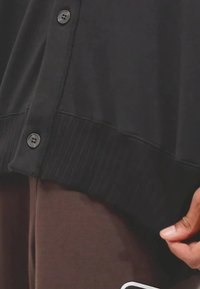 Sort cardigan med ribbet kant og fem knapper, vist nær brunt stof. Materialet ser blødt ud, med en glat tekstur og minimal udsmykning.