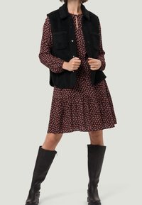 Schwarze Fleece-Weste mit zwei vorderen Taschen über einem dunkelblumigen Kleid. Knielange schwarze Lederstiefel runden das Outfit ab.