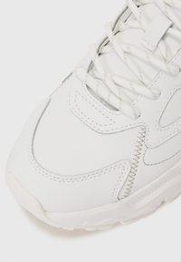 White sneaker with a smooth leather upper, stitching accents, and a textured sole. Features flat laces and a rounded toe design.