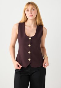 Bordeauxrode mouwloze gilet met een ronde halslijn, voorzien van vijf gestructureerde gouden knopen en een getailleerde pasvorm, gecombineerd met zwarte broek.