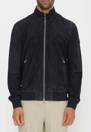 Uomo che indossa una giacca nera in suede con zip frontale, tasche con zip ai lati, polsini a coste e pantaloni beige su sfondo bianco.