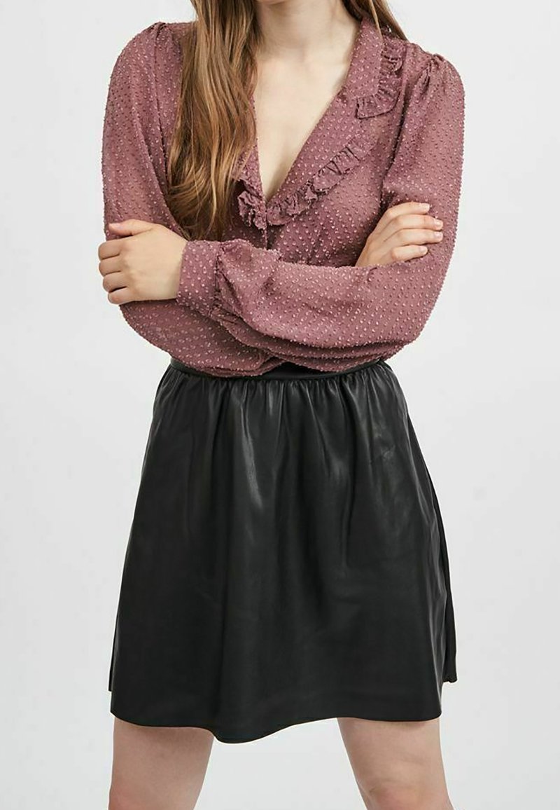 Femme portant un chemisier mauve texturé avec des volants et une jupe en cuir noire, debout les bras croisés devant un fond uni.