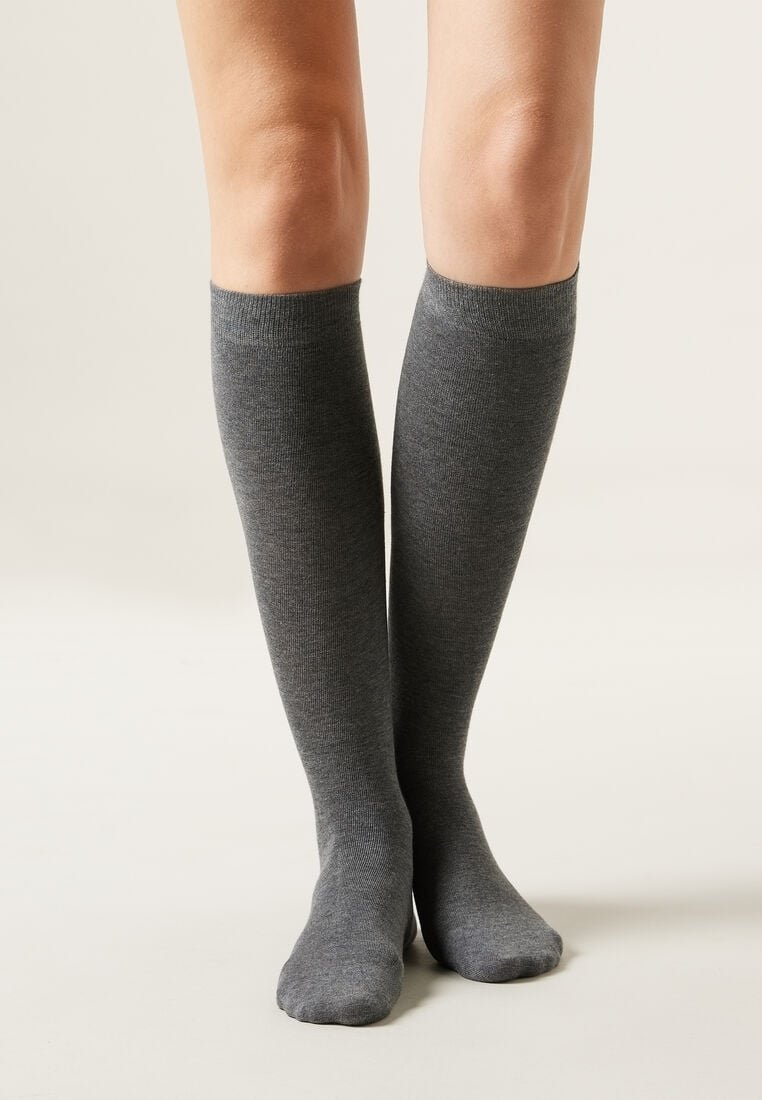 Jambes portant des chaussettes hautes gris foncé, debout sur un fond clair et neutre, les pieds rapprochés.