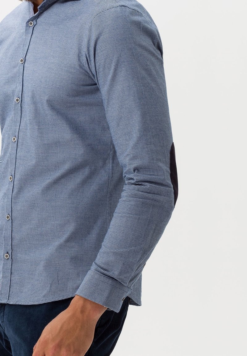 Homme portant une chemise bleue à motif micro-carreaux avec un patch au coude, associée à un pantalon bleu foncé sur fond blanc.