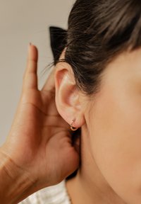 Pendiente de aro dorado con acentos de piedras preciosas coloridas, llevado en el lóbulo de la oreja, acompañado por una mano tocando la oreja.