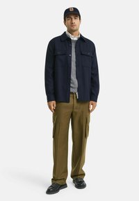 Marine knoop-overhemd jas over een grijze trui, donkerbruine cargobroek met zijzakken, en zwarte veterschoenen, afgewerkt met een marineblauwe pet.