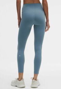 Leggings de talle alto y tejido elástico en azul claro, con textura suave y un logo sutil en el tobillo, combinados con zapatillas blancas.