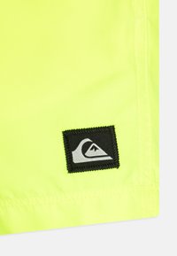 Neon sárga szövet sima textúrával; az alsó sarokban egy fekete folt található, rajta egy fehér hullám logóval.