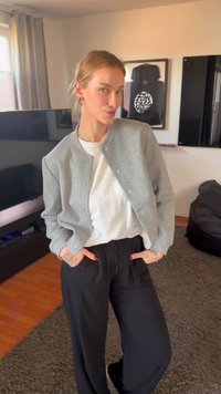 Mladá žena s blond vlasmi v sivom sakom, bielom tričku a čiernych nohaviciach stojí s rukami v vreckách v modernom obývacej izbe.