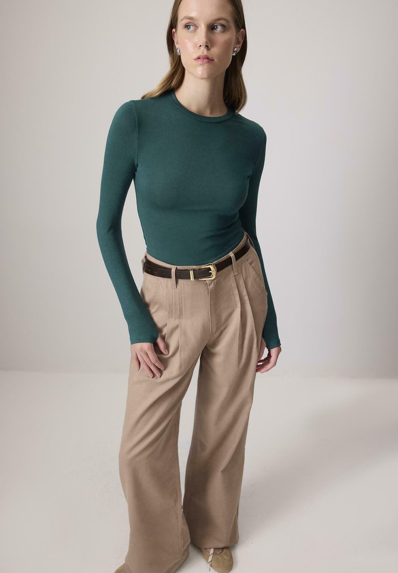 Naine seisab, seljas tumeroheline sobiv pikkade varrukatega särk, kõrge vöökohaga laiad beežid püksid tumeda vöödiga ja heledat pruunid kingad.