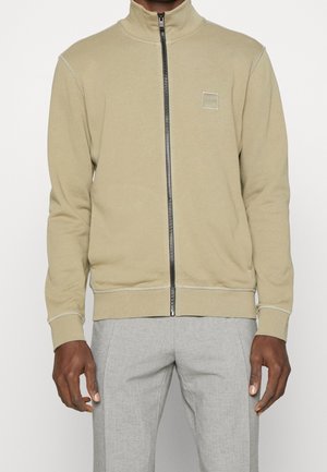 Mann trägt eine beige Jacke mit Reißverschluss und kleinem quadratischem Logo auf der Brust sowie hellgraue Hose, steht vor einem einfarbigen Hintergrund.
