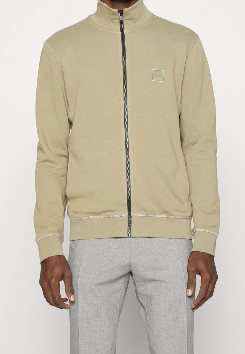 Homme portant une veste beige zippée avec un petit logo carré sur la poitrine et un pantalon gris clair, debout devant un fond uni.