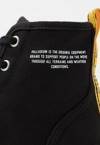 Bottine noire en toile avec accent jaune, dotée d'une fermeture à lacets et d'un texte imprimé détaillant le soutien de la marque pour tous les terrains et toutes les conditions météorologiques.