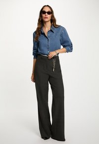 Camisa de mezclilla con adornos plateados, combinada con pantalones oscuros de pierna ancha. Los accesorios incluyen un cinturón de cadena y gafas de sol.