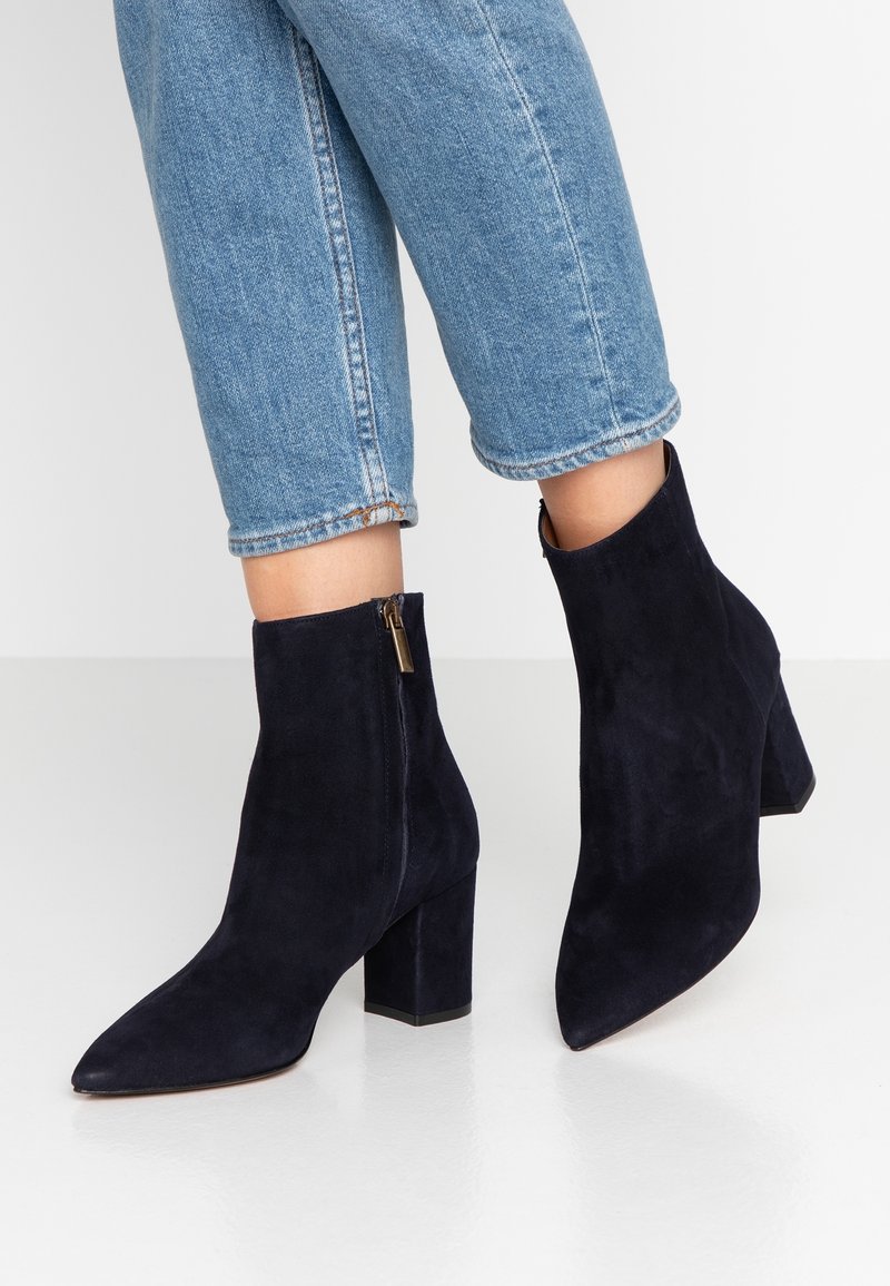 Femme portant un jean bleu court et des bottines en suède noir à talons carrés avec des fermetures éclair latérales, debout sur un sol blanc.