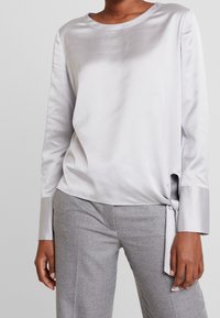 Zijdeachtige grijze top met lange mouwen en een ronde halslijn, zijden strikdetail en contrasterende manchetten. Gecombineerd met grijze, gestructureerde broek.