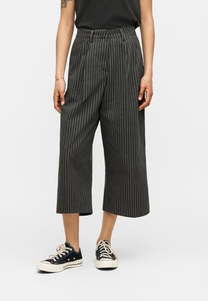 Personne portant un pantalon large court à fines rayures noires et blanches, un haut noir sans manches, des baskets noires et blanches à lacets, debout sur fond blanc.