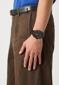 Pantalon marron avec une poche latérale, arborant des détails de couture bleus. Une montre-bracelet noire et marron avec un bracelet texturé repose sur la main.