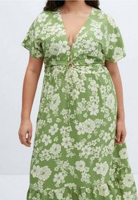 Femme portant une robe verte avec un motif floral blanc et un détail noué à l'avant, manches courtes et ourlet à volants.
