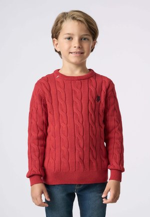 Maglione rosso a trecce con collo rotondo, polsini a coste e un piccolo logo sul petto. Indossato sopra a jeans blu.