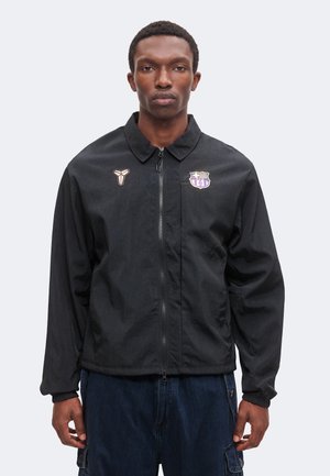 Mann trägt schwarze Jacke mit Reißverschluss und Sportlogos auf der Brust sowie dunkelblaue Cargohosen und steht vor einem schlichten hellen Hintergrund.