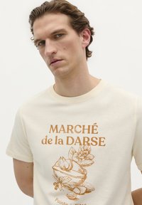 Crème katoenen T-shirt met een ronde halslijn, voorzien van een bruine grafische illustratie van een kom met bloemen en de tekst "MARCHÉ de la DARSE."