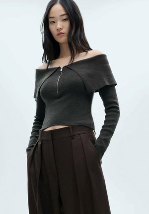 Donkergrijze off-the-shoulder top met een rits aan de voorkant, gecombineerd met hooggewaakte, geplooide bruine broeken. Zachte textuur, aansluitend ontwerp.