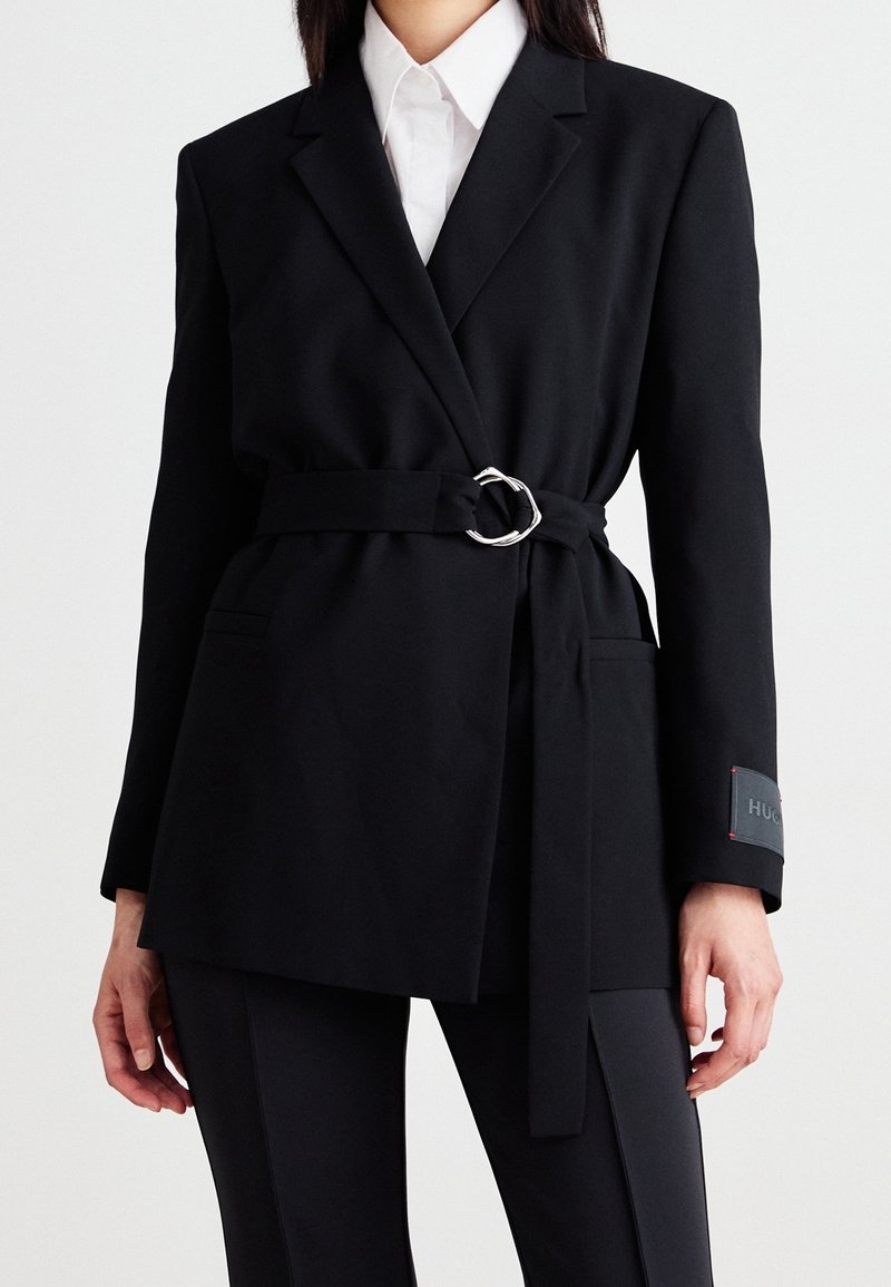 Femme portant un blazer noir ceinturé sur une chemise blanche à col et un pantalon noir, debout devant un fond blanc uni.