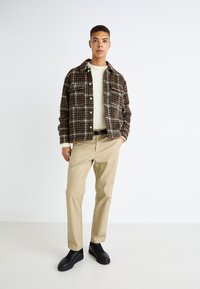 Braune karierte Shirtjacke, cremefarbener Pullover, beigefarbene Hose und schwarze Sneakers. Das Model steht in einem neutralen Raum mit den Händen an den Seiten.