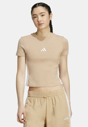 Giovane donna con capelli intrecciati indossa una t-shirt beige corta e pantaloni sportivi beige abbinati con strisce bianche e logo Adidas.