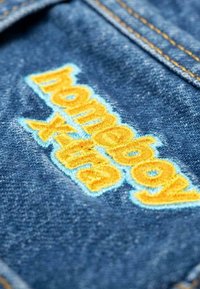 Denim materiale med blå tekstur; har gul og lys blå brodert tekst "homeboy xtra" tydelig vist.