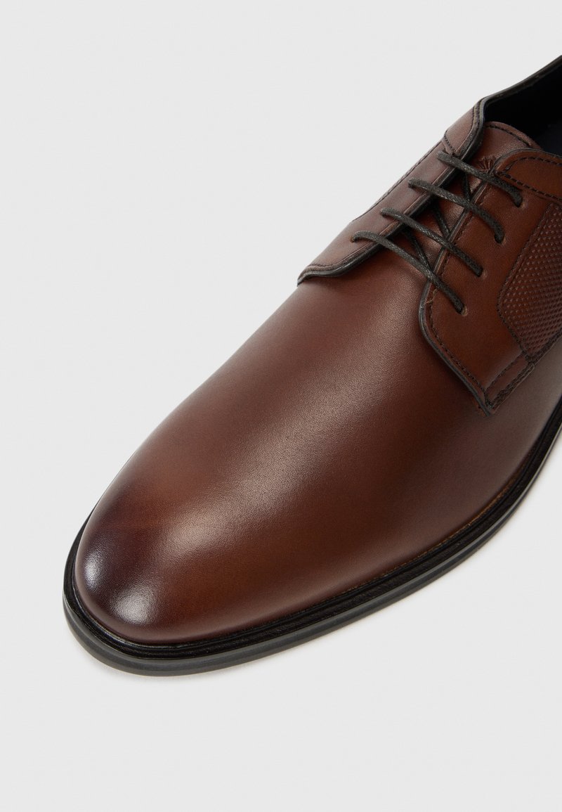 Brązowe skórzane męskie buty wizytowe z zamkniętym sznurowaniem, gładkim, zaokrąglonym noskiem i teksturowanymi bocznymi panelami na białym tle.