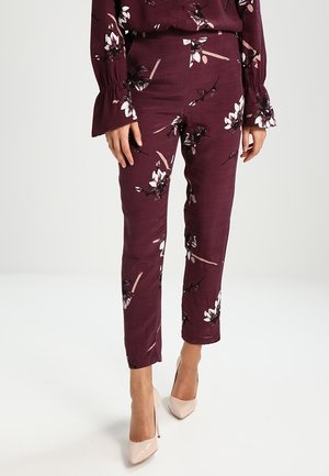 Pantalons imprimés floraux bordeaux avec une coupe slim, présentant des motifs de fleurs blanches et noires, fabriqués en tissu léger, associés à des talons beiges.