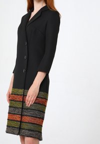 Svart kappa med en strukturerad silhuett, med tre knappar och en flerfärgad randig vävd kant i orange, grön och marinblå.