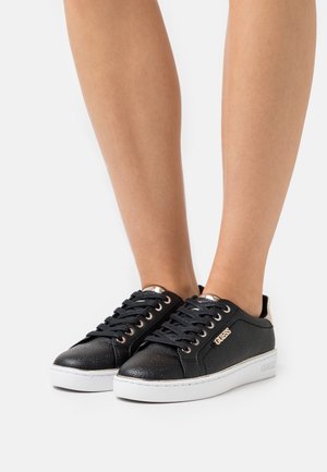 Baskets basses noires Guess avec motif embossé, logo doré sur le côté, semelles en caoutchouc blanches, portées pieds nus sur un fond uni.