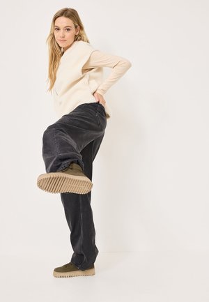 Gilet sans manches beige en polaire sur un top léger à manches longues. Pantalons larges noirs, bottines vertes avec des semelles texturées.
