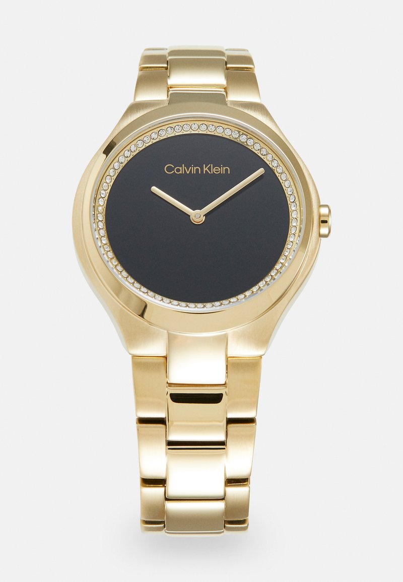 Reloj de tono dorado con una caja redonda, esfera negra y acentos de cristal alrededor del bisel. Presenta dos manecillas doradas y un estilo de pulsera eslabonada.