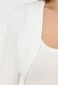 Femme portant un cardigan court blanc par-dessus un haut blanc à encolure dégagée, montrant l'épaule et le haut de la poitrine.