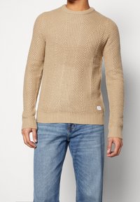 Vaurio beige neulepaita, jossa on teksturoitu kuvio, pyöreä kaula-aukko ja resorit hihansuissa. Yhdistetty sinisiin farkkuihin, joissa on pieni logo-tunnus.