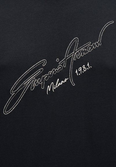 Μαύρο ύφασμα με λευκή κεντημένη γραφή που αναγράφει «Giorgio Armani Milano 1981» σε στυλ καλλιγραφικής γραφής.