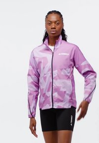 Lätt lila jacka med geometriska mönster, hel dragkedja, hög krage och svarta detaljer, bärs över åtsittande svarta shorts.