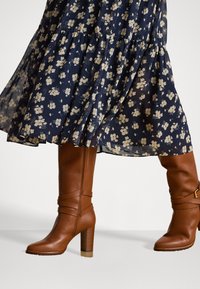 Donna che indossa stivali alti in pelle marrone e un vestito blu navy con stampa floreale beige, di lunghezza media e con dettagli in tessuto fluido.