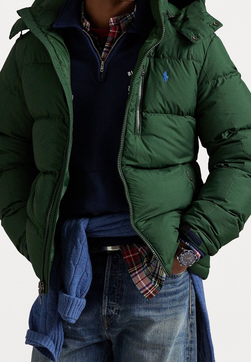 Personne portant une veste matelassée verte, une chemise à carreaux, un pull bleu marine, un pull bleu noué à la taille, un jean et une montre-bracelet.
