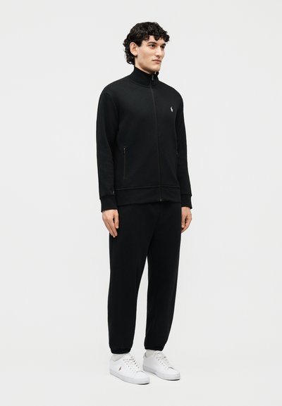 Mladý muž stojící, oblečený v černé bundě na zip a odpovídajících kalhotách s bílými teniskami na jednoduchém světlém pozadí.