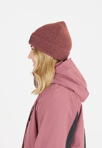 Ribattuto beanie in bordeaux, con un top appuntito e un'etichetta nera. Abbinato a una giacca con cappuccio rosa.