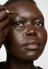 Nahaufnahme einer dunkelhäutigen Person, die einen braunen Augenbrauenstift aufträgt, mit glatter Haut, vollen Lippen und definierten Wangenknochen, vor einem schlichten Hintergrund.