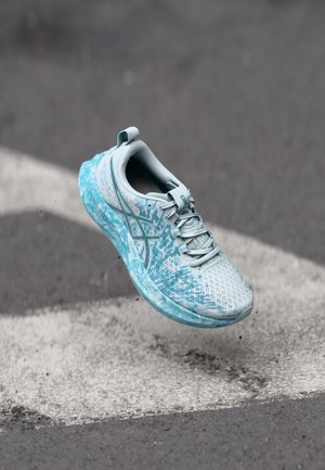 Light blue and white running shoe floating above asphalt with white road markings, small dirt particles visible beneath the shoe.
