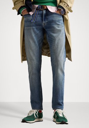 Mand iført blå jeans, grønne sneakers med hvide snørebånd, en beige trenchcoat og et blåt ur, hænderne i lommerne.